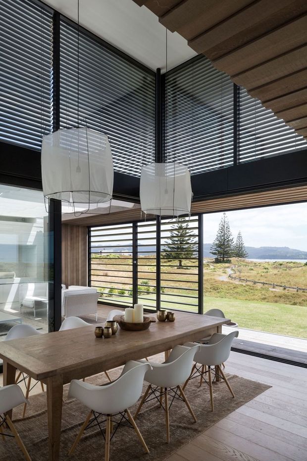 A contemporary dining area with a large timber dining table and white molded chairs, framed by extensive black aluminium glazing and horizontal blinds. Two oversized white pendant lights hang beneath a timber-lined ceiling, while large sliding doors open to a grassy coastal landscape and distant water views. The space has a warm, refined indoor-outdoor character with strong natural light and exposed structural framing.