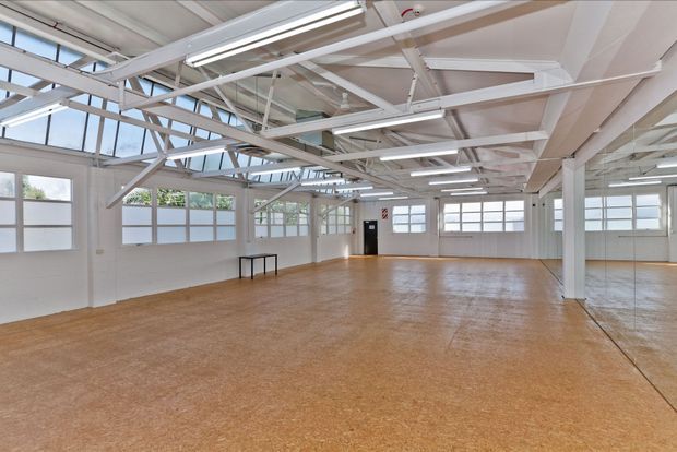 Large, bright open-plan commercial or industrial-style interior with a broad polished brown floor, white-painted structural beams and columns, extensive frosted and clear glazing, exposed roof framing, and linear fluorescent lighting. A full-height mirror on the right reflects the open space. The room is largely empty and suitable for studio, office, showroom, or flexible commercial use.