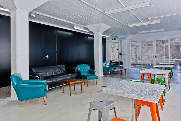 A bright open-plan commercial-style lounge and workspace with polished concrete flooring, exposed white-painted ceiling structure, black wall panelling, and large glazed doors and windows. Teal leather lounge chairs and sofas sit alongside small timber side tables, while colourful stools and communal tables occupy the foreground. The space has a contemporary industrial character with a high ceiling and abundant daylight.