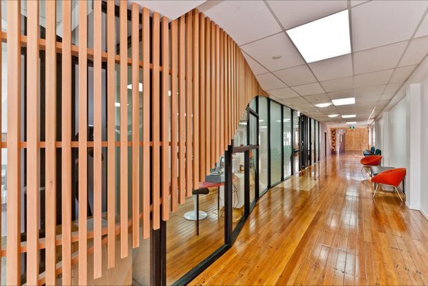 A bright contemporary commercial hallway with polished timber flooring, full-height glass office partitions in black frames, and a striking curved screen of vertical timber battens. The corridor includes modern red and orange lounge chairs, ceiling grid lighting, and glazed meeting or office spaces.