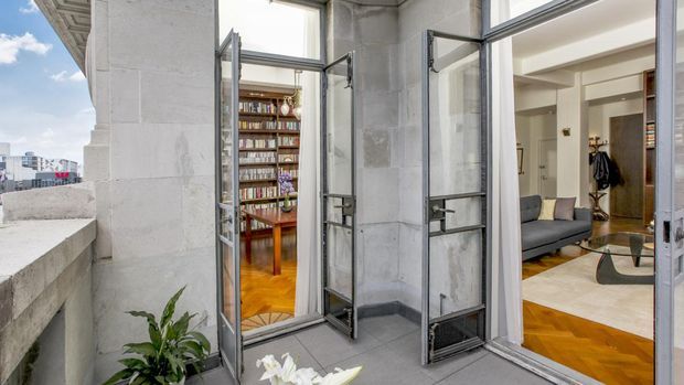 View from a balcony or terrace through open, black-framed glazed doors into a refined apartment interior. The foreground has pale stone or concrete walls and balustrade, while the interior beyond shows a light-toned living room with a dark sofa, herringbone timber flooring, white walls, and a bookshelf-lined room visible through another opening. Urban buildings are visible in the distance.