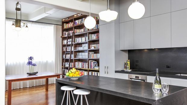 A bright contemporary open-plan kitchen and dining area. The kitchen features a large dark stone island with white stools, extensive matte grey cabinetry, a dark backsplash, integrated appliances, and white pendant lights. Beyond the island is a timber dining table beside a bright curtained window, with a full-height dark timber bookshelf forming a prominent feature wall.