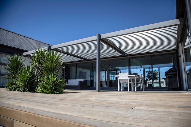 Contemporary single-storey home exterior featuring a broad timber deck, a covered outdoor entertaining area with a slatted pergola roof, dark-framed floor-to-ceiling glazing, outdoor dining furniture, and a built-in barbecue. Sculptural spiky planting borders the deck, while corrugated metal roofing is visible on the adjoining structure.
