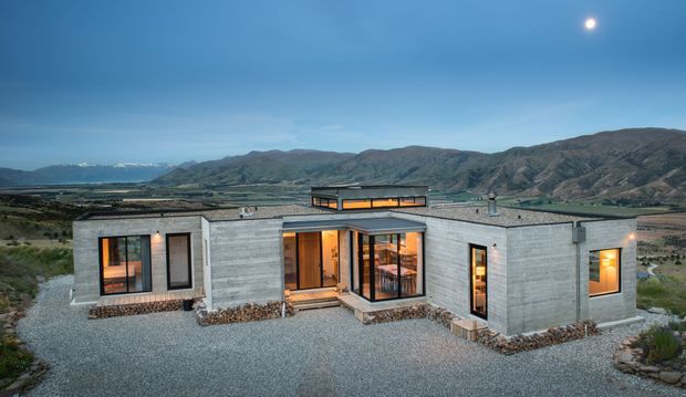 A low-profile modern concrete house set in a rugged mountainous landscape, photographed at dusk. The single-storey dwelling has a rectilinear, courtyard-like plan with exposed grey concrete walls, extensive dark-framed glazing, warm interior lighting, a raised central roof element or clerestory, and gravel landscaping with stone-edged planting beds. Mountains and a broad rural valley form the background, with the moon visible in the blue evening sky.