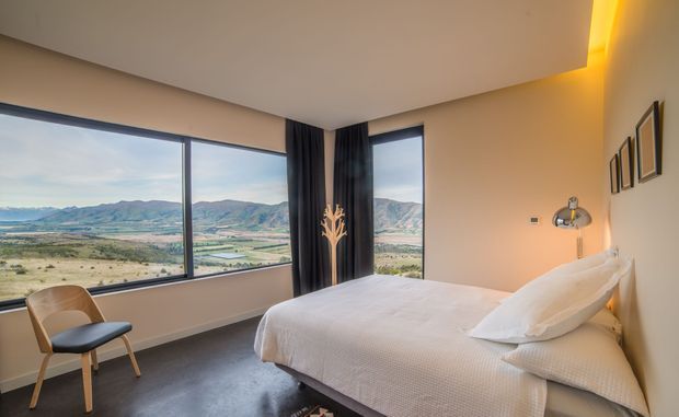 A serene modern bedroom with a large bed positioned beside expansive corner glazing, offering panoramic views of a rural mountainous landscape. The room features warm beige walls, dark floor finishes, floor-to-ceiling dark curtains, minimalist framed wall art, a sculptural bedside lamp, and concealed warm cove lighting along the ceiling edge.