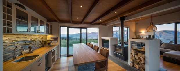 Open-plan kitchen and dining area with a long timber dining table, extensive built-in cabinetry, stone-clad splashback, and a compact fireplace dividing the dining zone from a living area. Exposed timber ceiling beams and large glazed doors frame a mountain and valley outlook. The space has a warm, refined alpine character with layered timber and stone finishes.