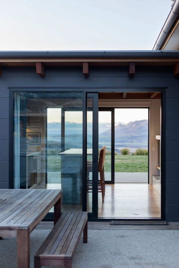 View from an outdoor deck toward a dark-clad contemporary house, with timber picnic-style bench seating in the foreground and large sliding glass doors opening into a bright dining and living area. The interior has polished light flooring and timber ceiling detailing, while a dramatic lake, grassland, and mountain landscape is visible through the glazing.