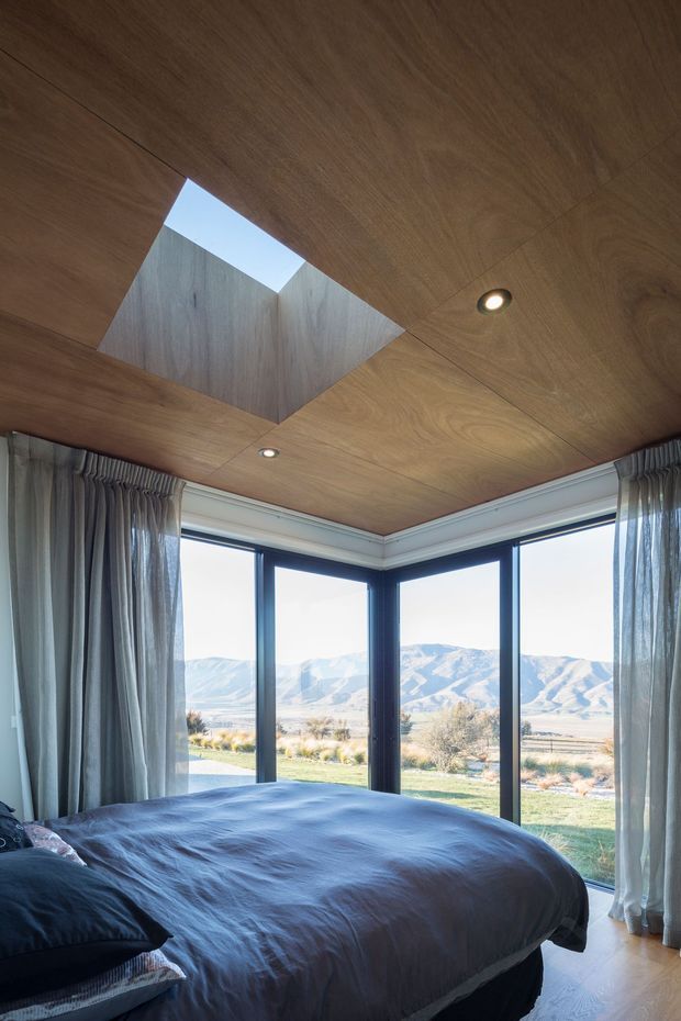 A contemporary bedroom with a low warm timber-lined ceiling, recessed downlights, and a dramatic square skylight opening. Floor-to-ceiling corner glazing with dark frames and sheer curtains overlooks a sunlit mountainous landscape. A large bed with dark blue-grey bedding occupies the foreground, creating a calm, nature-connected atmosphere.