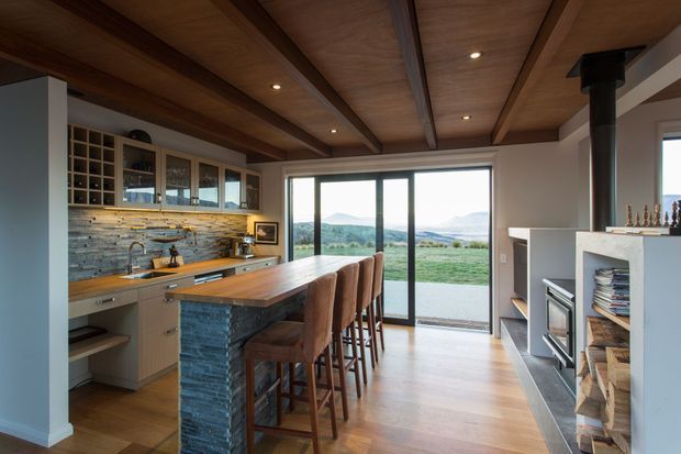 A warm open-plan kitchen and living area with a long timber island, four upholstered bar stools, built-in cabinetry, a natural stone island base, and a rustic stone backsplash. The room features a timber-lined ceiling with exposed dark beams, timber flooring, large glazed sliding doors opening to a rural landscape, and a contemporary fireplace with integrated shelving and firewood storage.