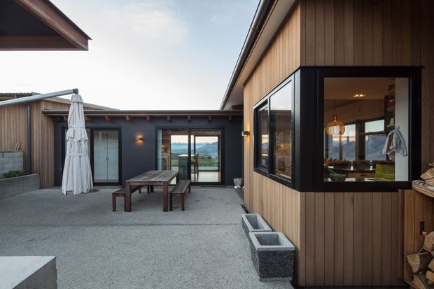 A contemporary timber-clad courtyard or terrace framed by low-pitched roof forms. The outdoor space has a textured grey paved surface, a timber picnic-style dining table with benches, a closed outdoor umbrella, wall-mounted lighting, planter boxes, and large glazed openings. Through the glazing, a dining area and scenic landscape are visible. The composition emphasizes a private, sheltered courtyard connected to the interior.