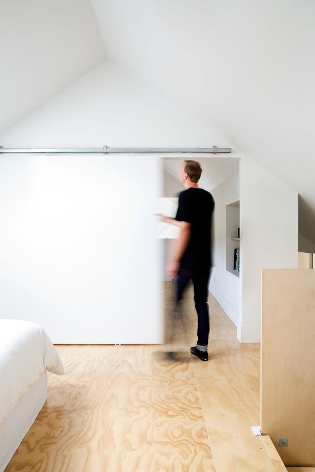 Bright minimalist interior viewed from a bedroom or sleeping area, featuring pale plywood flooring, white walls, a large white sliding barn-style door on exposed metal hardware, and a compact hallway or storage zone beyond. A person is walking through the space, with a partial bed visible at lower left and built-in shelving recessed into the wall.