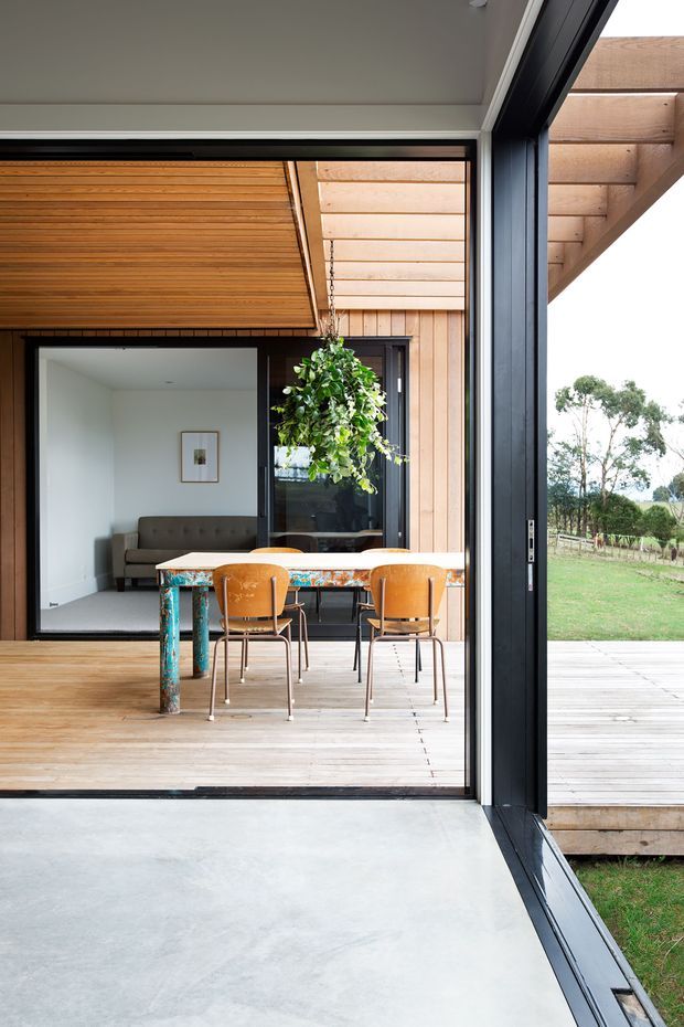 A contemporary indoor-outdoor dining area opening onto a timber deck through large black-framed sliding or folding glass doors. The space features a timber-lined ceiling and wall, polished concrete flooring inside, a rustic timber dining table with colourful legs, and simple wooden dining chairs. A compact living area is visible beyond, with landscaped rural surroundings outside.