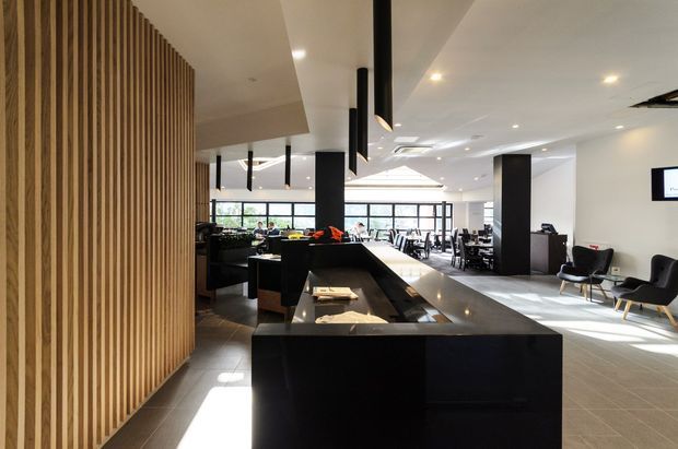 A spacious contemporary commercial interior, likely a hospitality or office reception and lounge area. The room features a long black stone-clad counter or island in the foreground, a full-height vertical timber slat feature wall on the left, black structural columns, suspended black pendant lights, tiled flooring, extensive glazing, and scattered tables and lounge seating in the background. The palette is neutral with black, white, grey, and warm timber accents, creating a refined and bright atmosphere.