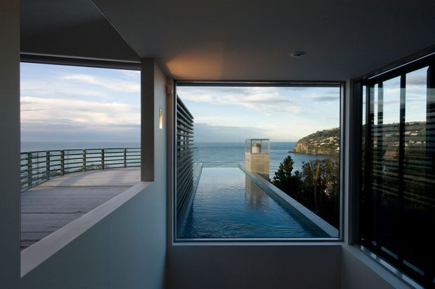 Interior view from a modern coastal residence looking through large floor-to-ceiling windows toward the ocean, a rocky headland, and a projecting balcony or deck. The space has a dark ceiling, smooth pale walls, slim dark window frames, and a strong indoor-outdoor connection. A timber-toned deck and metal balustrade are visible at left, while reflective glazing and the ocean dominate the view.