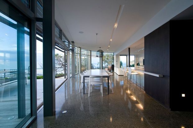 A spacious contemporary open-plan kitchen and dining area with floor-to-ceiling glazing along one side, opening to an outdoor deck and expansive water views. The room features a long dining table, bar stools at a kitchen island, dark full-height cabinetry, integrated lighting, and a polished speckled hard floor. Strong indoor–outdoor connection and abundant natural light define the space.