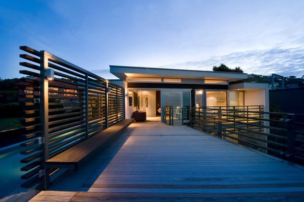 Twilight exterior view of a contemporary flat-roofed home accessed via a broad timber deck or rooftop terrace. Horizontal dark metal screens and balustrades frame the approach, while warm interior lighting glows through expansive glazing. The image emphasizes the elevated outdoor circulation space, clean geometric lines, and strong indoor-outdoor connection.