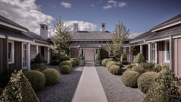 A symmetrical landscaped courtyard framed by low-rise wings of a refined residential or hospitality-style building. A straight pale paved walkway leads to a central entrance, with dark gravel beds, clipped spherical shrubs, conical evergreens, young trees, and muted timber-clad façades. The buildings have pitched shingle roofs, white-trimmed windows and doors, and multiple chimney-like roof projections under a dramatic partly cloudy sky.