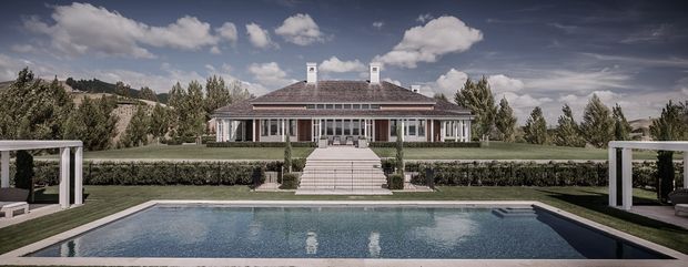 A symmetrical exterior view of a large single-storey residence set in a landscaped rural or suburban site, with a broad hipped roof, dormer-like chimney structures, classical columns and evenly spaced windows. A central staircase leads from the lawn to a covered veranda and the house entrance, while a rectangular swimming pool and paved terrace occupy the foreground. White-framed pavilion structures sit on both sides of the pool. The image has a cinematic, slightly muted presentation with dramatic clouds and a formal, refined composition.