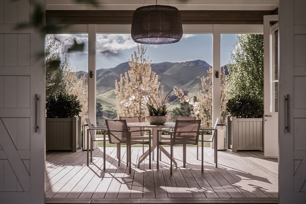 Bright, refined dining area opening onto a mountain landscape through large glazed doors. A rectangular dining table with four slim-framed chairs sits on a pale timber deck or floor, flanked by built-in white cabinetry and tall planters. A woven pendant light hangs centrally, while soft neutral finishes and abundant daylight create a calm indoor-outdoor atmosphere.