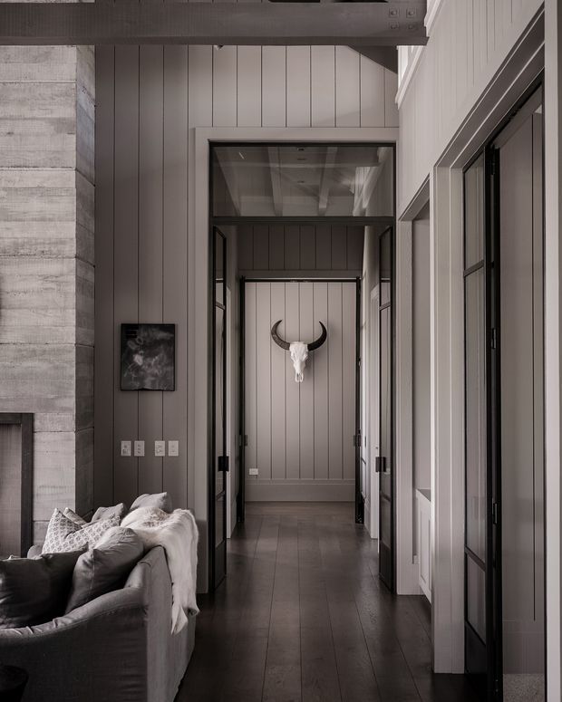 A refined interior view from a living room toward a tall, paneled hallway or entry passage. The space features dark timber flooring, vertically grooved grey wall panelling, white trim and doors, black-framed glazed internal doors, exposed dark ceiling beams, and a decorative cow skull mounted at the end of the corridor. A portion of a large upholstered sofa is visible in the foreground.