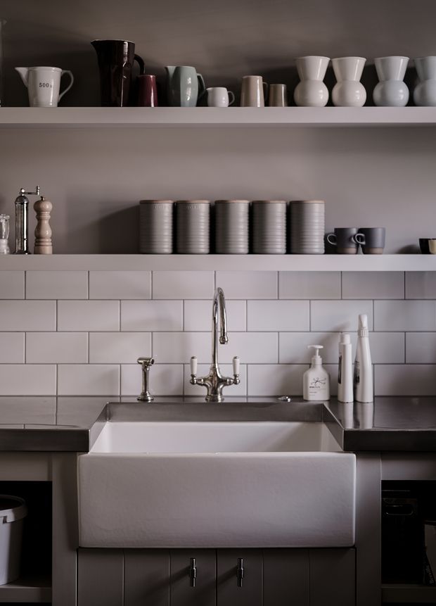 Close-up view of a refined kitchen sink area with a large white apron-front farmhouse sink, polished metal bridge-style tapware, and white rectangular subway-tile splashback. Open shelving above displays ceramic pitchers, vases, cups, and ribbed storage canisters. Dark benchtops and cabinetry contrast with the light tiled wall and white sink, creating a classic, softly moody kitchen composition.