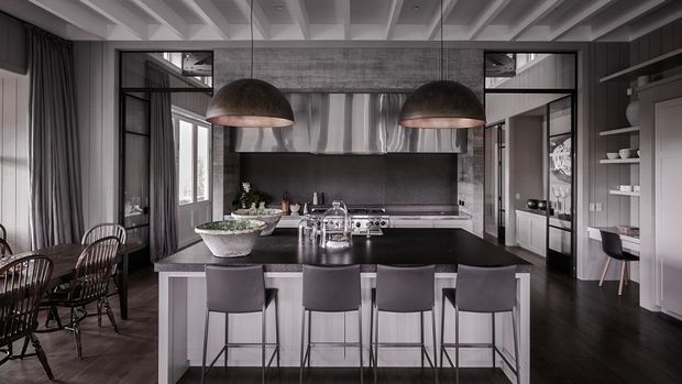 A sophisticated open-plan kitchen and dining area in a dark monochromatic palette. The kitchen features a large central island with four bar stools, dark stone-like benchtops, pale cabinetry, integrated appliances, a stainless-steel rangehood, and two oversized dome pendant lights. The room includes a dining table at left, dark timber flooring, expansive steel-framed glazing and doors, open shelving, and a distinctive coffered ceiling with exposed beam detailing.