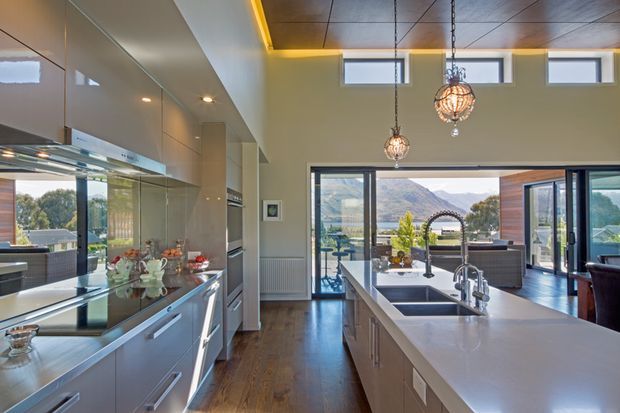 A contemporary open-plan kitchen with a long central island, white integrated cabinetry, dark timber flooring, stainless-steel appliances and a deep double sink. Large glazed sliding doors open to a terrace or outdoor living area with a mountain and water view. High clerestory windows, a timber-toned ceiling, concealed cove lighting and hanging globe pendants add architectural interest.