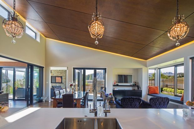 Open-plan kitchen and dining/living area with a broad pale benchtop and inset stainless-steel sink in the foreground. The room has a dark timber-panel ceiling with warm concealed perimeter lighting, pale walls, large glazed doors and windows, a dining table, patterned pendant lights, and a separate lounge area with a wall-mounted television and fireplace or media joinery. Outdoor views are visible through the glazing.