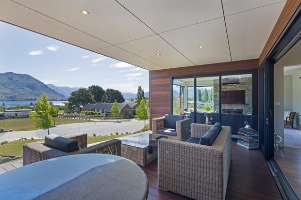 A covered outdoor terrace overlooking a landscaped residential setting and mountain-and-lake landscape. The space features dark timber decking, warm horizontal timber wall cladding, a pale panelled ceiling with recessed downlights, extensive black-framed glazing and sliding doors, and a seating arrangement of woven outdoor armchairs and sofas around a low coffee table. An interior living space is visible through the open glazing.