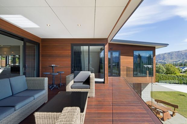 Contemporary covered outdoor terrace adjoining a wood-clad home, furnished with woven lounge seating, a dark coffee table and a small bar-height table. Timber decking extends beside floor-to-ceiling glazing and a glass balustrade, with mountain scenery and landscaped grounds beyond.