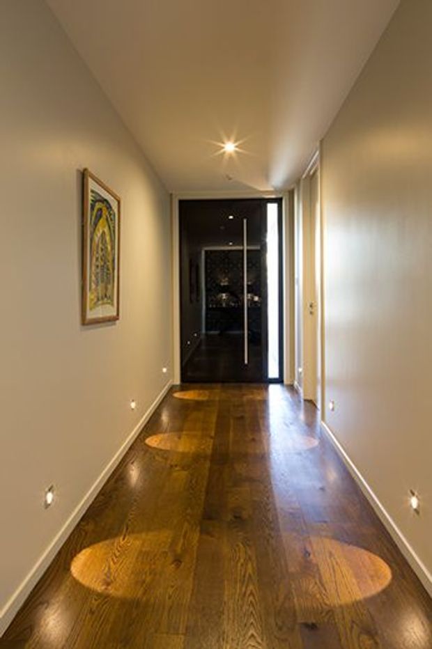 A narrow, elongated residential hallway with warm beige painted walls, a polished dark timber floor, recessed low-level wall lights, and a ceiling downlight. A framed artwork hangs on the left wall, while a dark glass or timber door with a long vertical handle terminates the corridor. Bright daylight enters through glazing or openings along the right side, creating circular reflections on the floor.