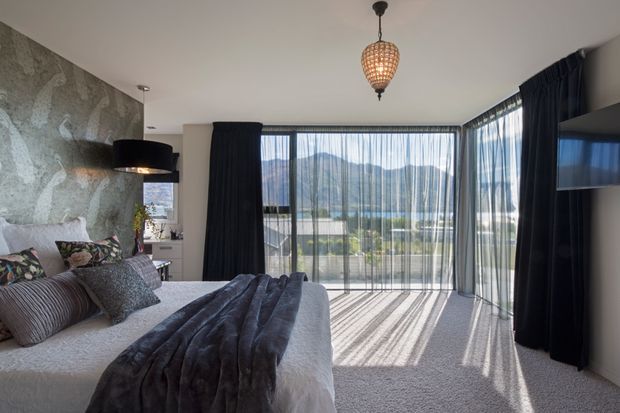 A contemporary bedroom with a large bed, textured grey bedding, patterned cushions, a dark suspended bedside light, and a sculptural textured feature wall. Full-height glazed doors and sheer curtains open to a balcony or terrace with expansive mountain and landscape views. Dark blackout curtains and a decorative pendant add contrast and warmth.