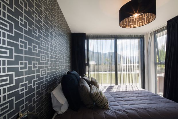 A stylish bedroom with a low bed against a dramatic dark feature wall covered in a geometric linear pattern. Floor-to-ceiling glazed doors and windows provide abundant daylight, with sheer curtains and dark drapes framing views of the outdoors. The room is finished with layered dark bedding, patterned cushions, and a large black pendant light.