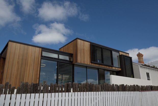 Exterior view of a contemporary multi-level residence with warm vertical timber cladding, extensive dark-framed glazing, and black louvred sun-shading panels. The home has a varied pitched roofline and sits behind a white slatted fence, with an older weatherboard dwelling visible alongside it.