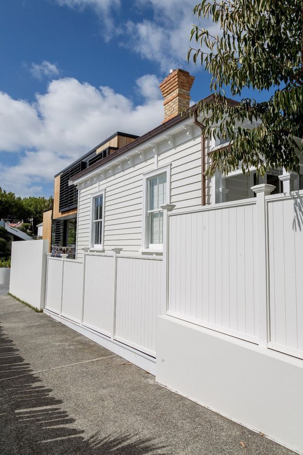 Exterior view of a white weatherboard house with a red-brick chimney, traditional sash-style windows, decorative eaves, and a tall white privacy fence. A contemporary dark-clad addition with horizontal screening is visible behind the original dwelling. The house fronts a concrete-paved walkway under a bright blue sky.