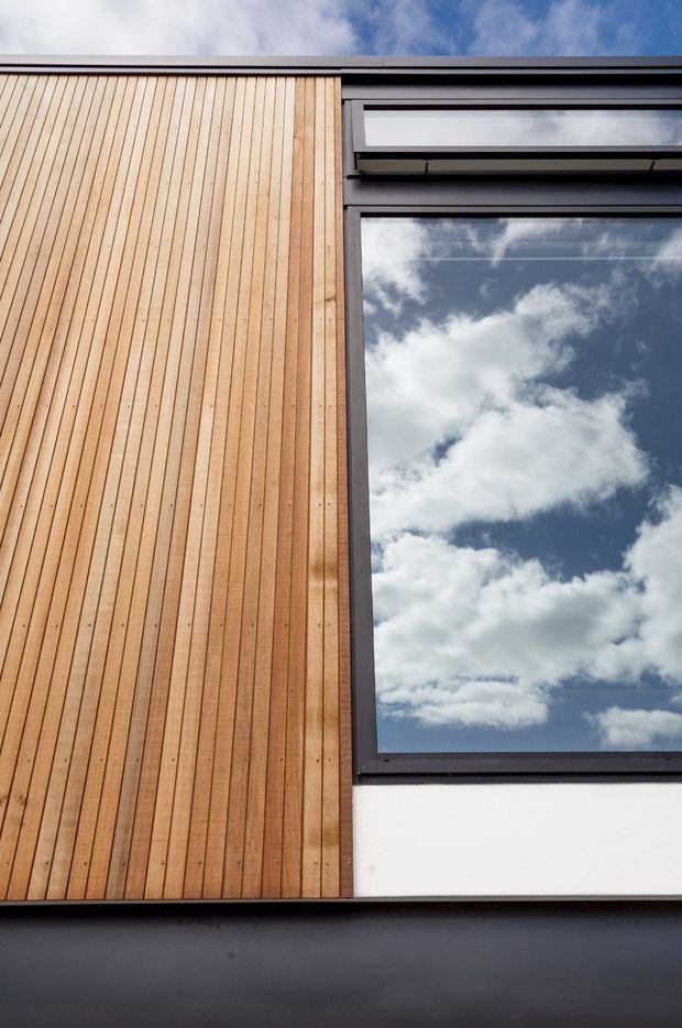 Close-up exterior architectural detail showing vertical warm timber cladding beside a large dark-framed window reflecting a partly cloudy sky. A crisp white wall panel and dark horizontal roof or sill elements create a strongly contrasting contemporary facade composition.