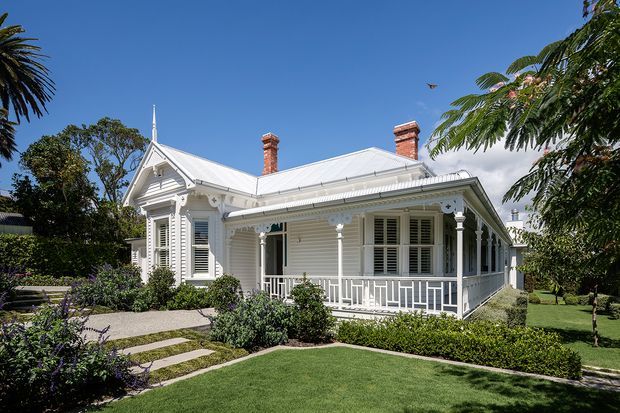 Exterior view of a restored single-storey Victorian-era cottage with a light weatherboard facade, ornate white timber trims, decorative gable detailing, louvred shutters, a broad covered veranda, brick chimneys and a steep metal roof with a small finial. The house is set within a manicured suburban garden with clipped hedges, lawn and a stepping-stone path.