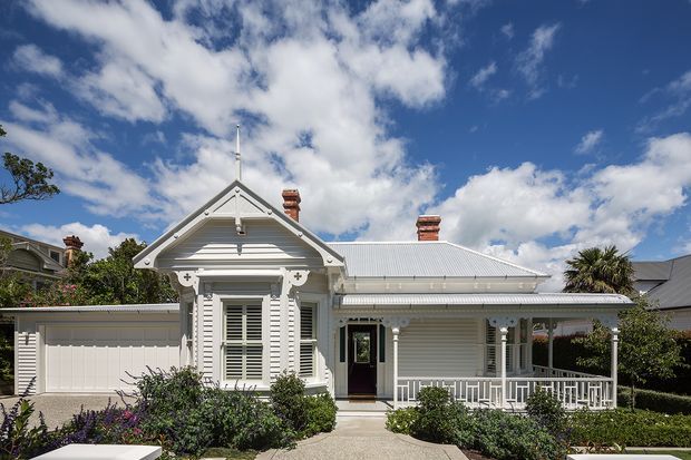 Street-facing view of a single-storey white weatherboard house with a steep gabled front projection, ornate decorative trims, louvred shutters, a corrugated metal roof, covered verandah, attached garage, and landscaped garden. The bright exterior is photographed under a vivid blue sky.