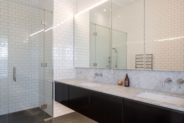 Bright contemporary bathroom with white subway-tiled walls, a glass-enclosed walk-in shower, expansive mirrored wall cabinetry, and a long double vanity. The vanity features dark flat-panel cabinetry, a pale marble benchtop and matching marble splashback, with chrome tapware and a heated towel rail. Small-format dark floor tiles provide contrast.
