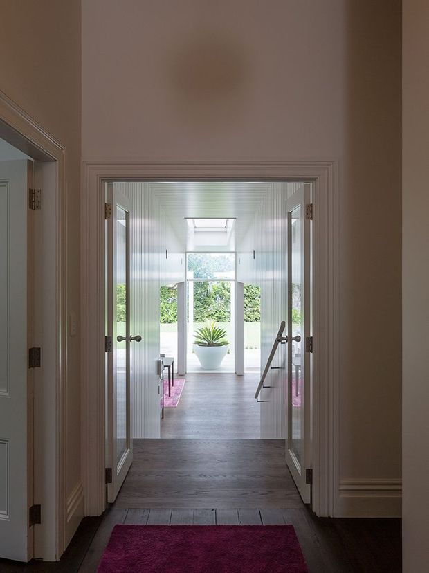 View through a traditional, heavily trimmed hallway into a bright glazed transition space or sunroom. Double doors open onto a room lined with white vertical wall panelling, dark timber flooring, a central skylight, large windows, and a potted plant visible beyond. A magenta runner adds colour in the foreground, while the surrounding hall has warm taupe walls and detailed mouldings.