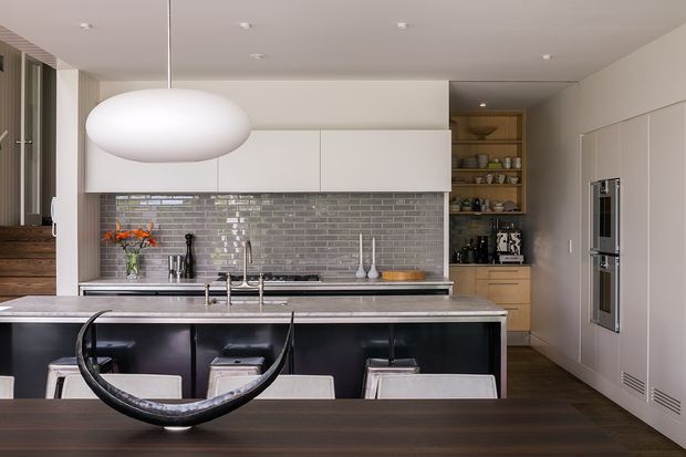 A refined contemporary kitchen viewed from the dining area, featuring a large island with dark glossy cabinetry and a stone benchtop, a grey brick-look tiled splashback, white overhead cabinetry, integrated appliances, open timber shelving, and a sculptural pendant light. Dark timber flooring and restrained neutral finishes create a polished, modern atmosphere.