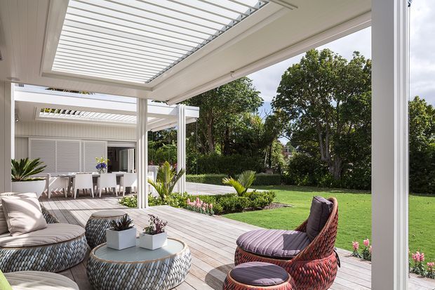 A bright covered outdoor entertaining area opening onto a landscaped garden. The space features a timber decking floor, white structural posts and beams, a louvred pergola roof, dining setting, woven lounge furniture, potted plants, and a manicured lawn with garden beds beyond.