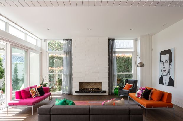 Bright, spacious contemporary living room with floor-to-ceiling glazing, a white painted brick fireplace wall, vertical white wall panelling, and a long low charcoal sectional sofa. Vibrant pink and orange lounge chairs with colourful cushions add playful contrast, while sheer curtains, a sculptural pendant light, and a large monochrome portrait complete the refined modern interior.