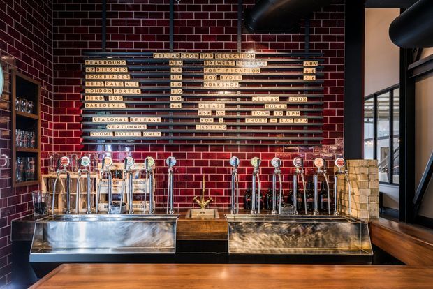 A commercial bar service area with a long polished timber counter, stainless-steel beer drip trays, numerous beer taps, and a dark red glazed brick feature wall. Timber-framed shelving with bottles is visible on the left, while black exposed ductwork and steel structural elements frame the space. A vintage-style letter board menu is mounted above the taps, and large windows bring in daylight at the right.
