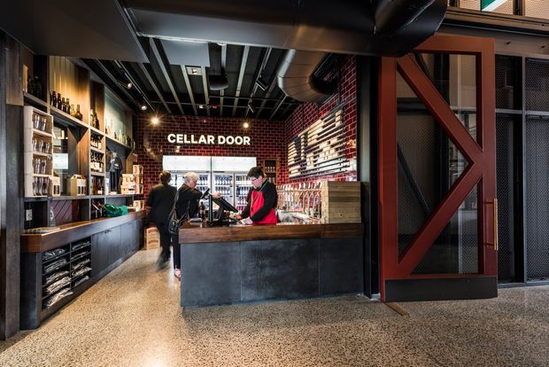 Interior view of a compact wine bar or cellar-door retail space. A dark service counter occupies the foreground, with extensive bottle shelving and display joinery along the walls. The space has exposed black ceiling services and ductwork, red brick walls, timber shelving, black metal framing, and a large industrial-style timber-braced door. Staff and customers are present behind and around the counter.