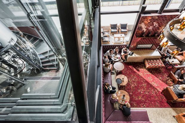 High-angle view into a large industrial-style commercial lounge or bar, with exposed metal services and structural elements, extensive glazing, a red brick feature wall, curved upholstered banquette seating, scattered round tables, a large patterned rug, and a bar counter. The left side shows a dramatic glazed view of exposed building infrastructure and ductwork, emphasizing an adaptive industrial interior.