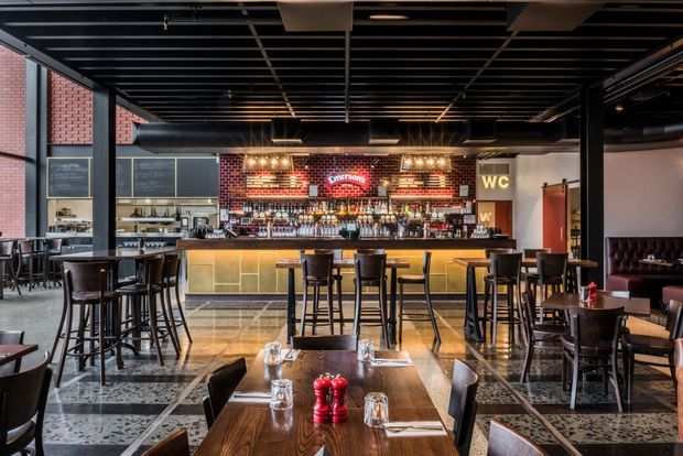 A spacious industrial-style bar and dining venue with a long illuminated bar counter, extensive bottle display, high stools, and numerous dark timber dining tables and chairs. Exposed black ceiling services and beams, steel-framed glazing, red brick walls, pendant and strip lighting, and patterned tiled flooring define the commercial interior. A small lounge booth is visible at the right.
