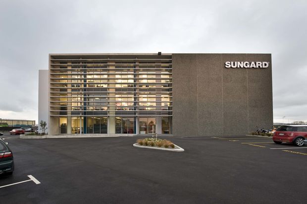 Exterior view of a contemporary commercial building with a large glazed entrance screened by horizontal fins, a tall dark textured facade panel, and prominent SUNGARD signage. The building fronts a paved parking area under an overcast sky.