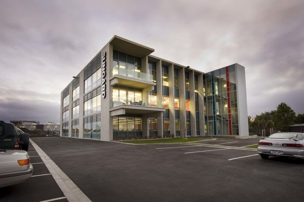Exterior view of a contemporary multi-storey commercial building with a rectilinear form, extensive glazing, vertical metal fins, projecting balconies or canopies, and a tall glazed feature volume with red and grey vertical panels. The building fronts a marked asphalt parking area under an overcast sky.