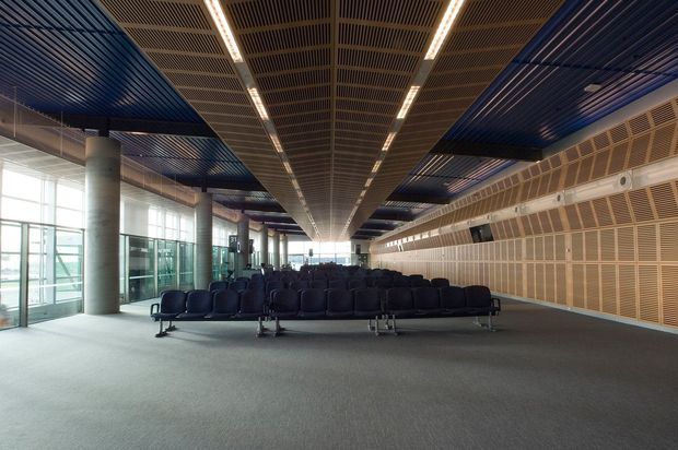 Large modern airport or transport terminal waiting area with rows of dark upholstered seating, expansive floor-to-ceiling glazing on the left, a long perforated acoustic wall on the right, cylindrical concrete columns, and a dramatic suspended linear ceiling with blue and timber-toned acoustic panels and integrated strip lighting.